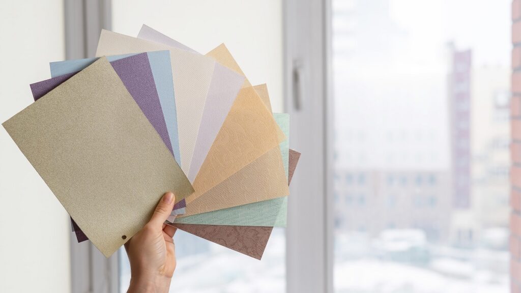 Woman holding fabric samples of roller blinds against window background.